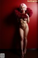 A nude woman with pink hair posing in front of a red wall.